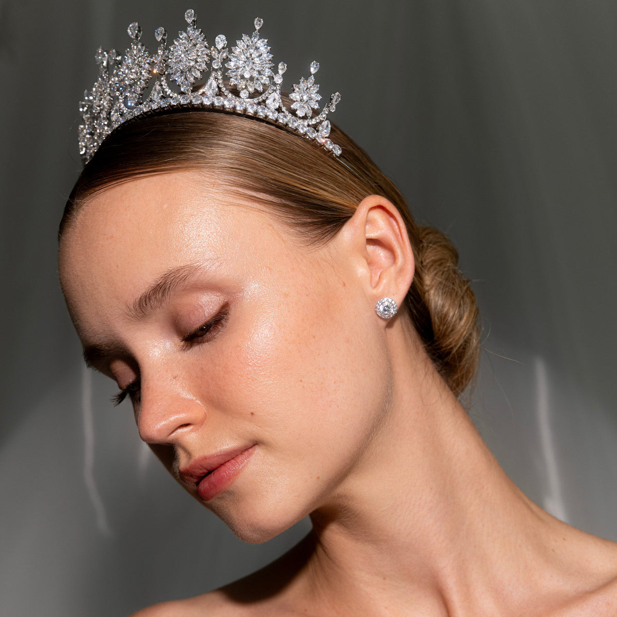 A woman wearing a sparkling silver tiara with clear stones and a matching stud earring, photographed in soft studio lighting against a dark gray background.
